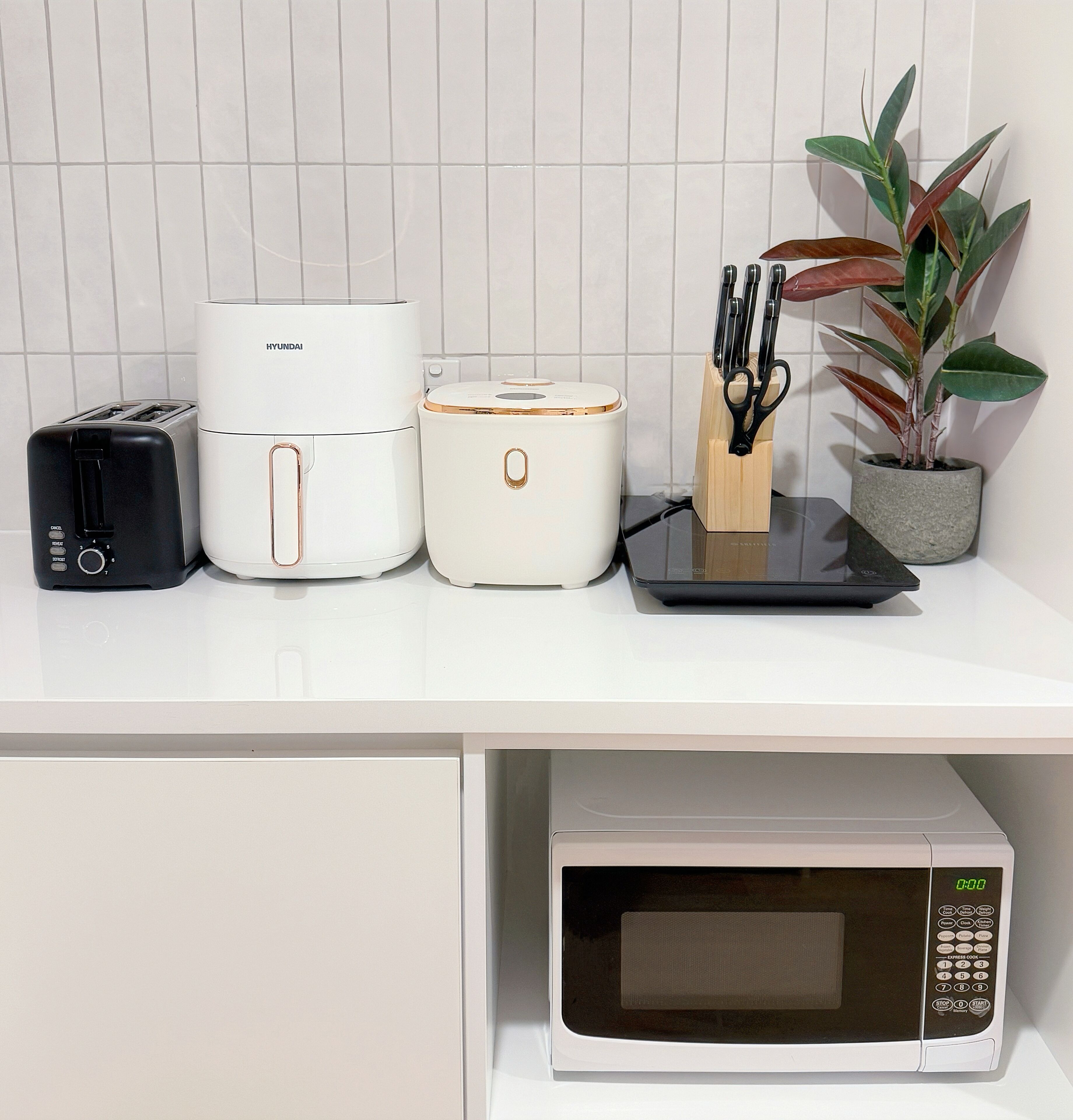 A kitchen counter with a toaster, rice cooker, microwave, and a plant.