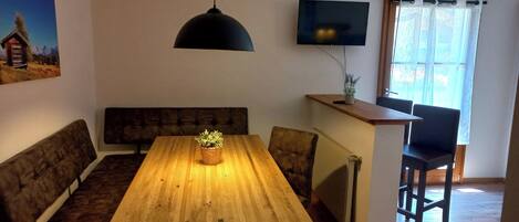 A dining area with a wooden table, benches, a hanging light fixture, a television on the wall, and a window with curtains.