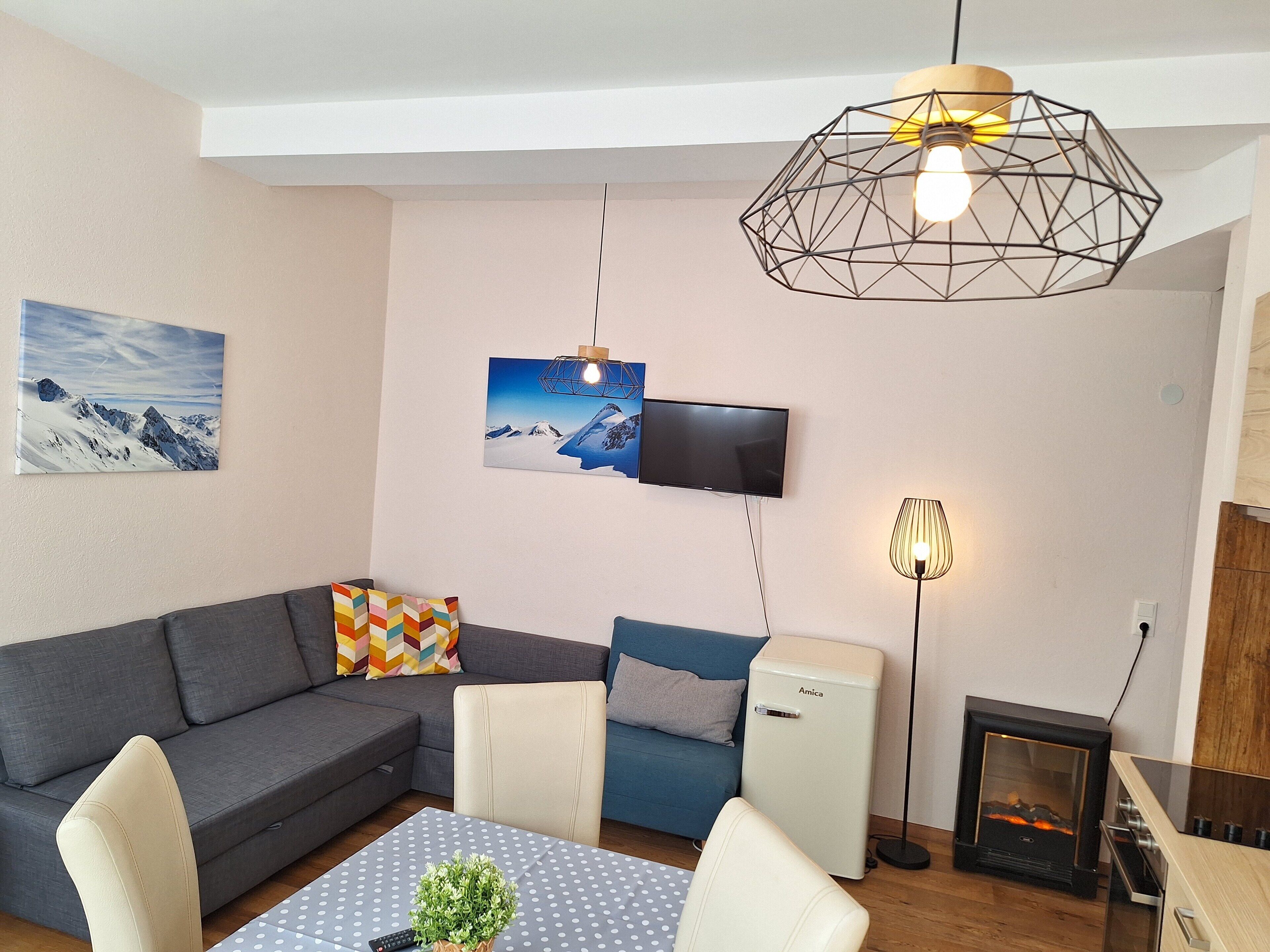 Un salon moderne avec un canapé gris, un fauteuil bleu, une petite table recouverte d’une nappe à pois, un téléviseur fixé au mur et un poêle à bois compact.