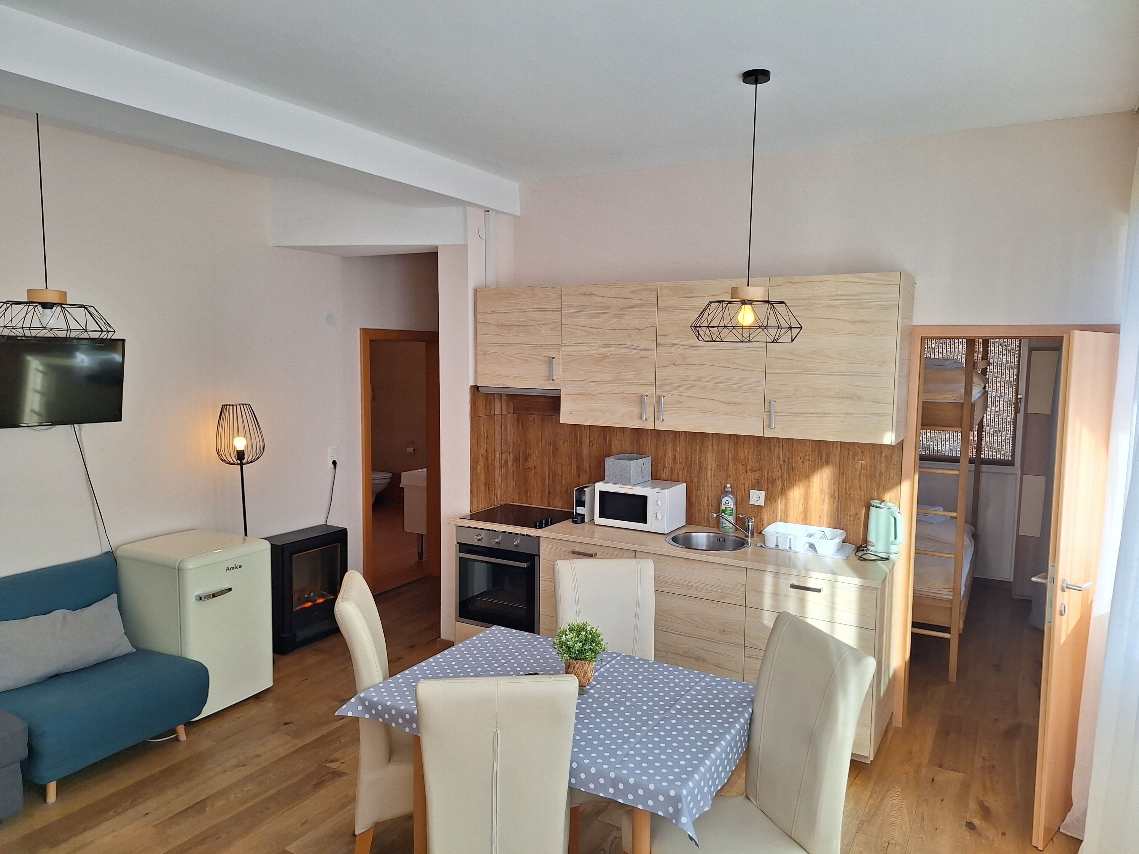 A compact kitchen with wooden cabinets, a microwave, and a sink. There is a dining table with chairs and a small plant. A sofa and a TV are also visible.