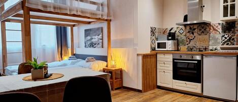 Una habitación de hotel moderna con una cama nido de madera, una zona de comedor con mesa y sillas, una cocina con armarios blancos y un microondas, y vistas a las montañas desde la ventana.