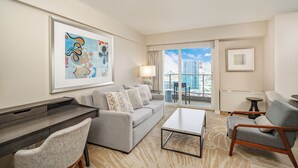 A modern hotel room with a grey sofa, a coffee table, and a desk. There is a large window with a view of buildings. - Junior Suite 1 Bedroom Ala Moana Condominium, City / Partial Ocean from Lanai (Honolulu)