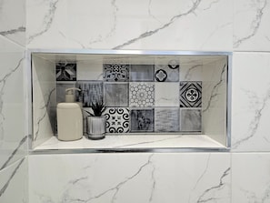A bathroom with a tiled backsplash featuring various patterns and a small shelf with a plant and a container.