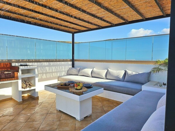 A rooftop terrace with a white sofa, a glass coffee table, and a barbecue grill.