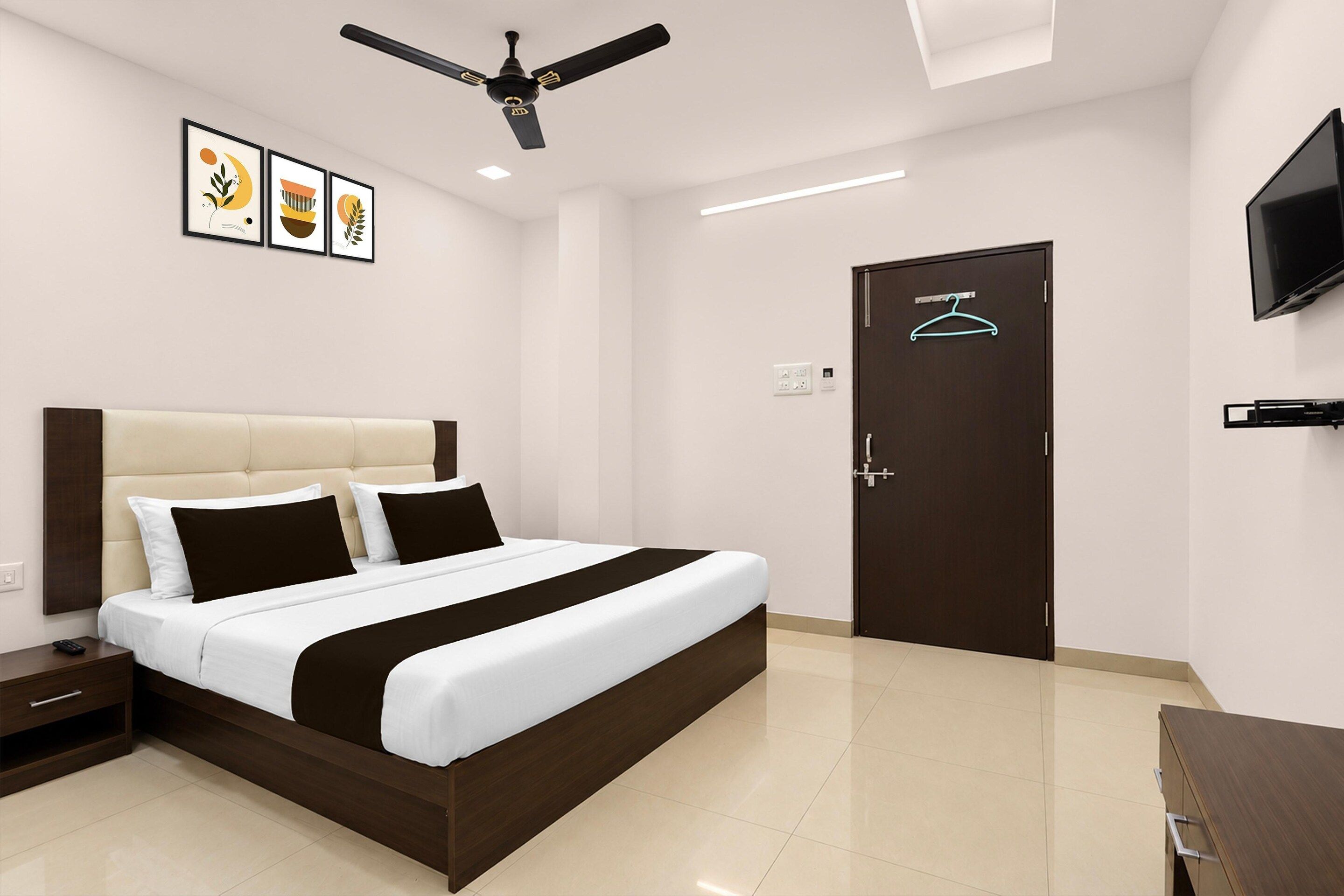 A hotel room with a bed, bedside table, nightstand, ceiling fan, wall-mounted TV, and a door with a hanger.
