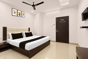 Habitación de hotel con cama, mesita de noche, ventilador de techo, televisor montado en la pared y una puerta con perchero.