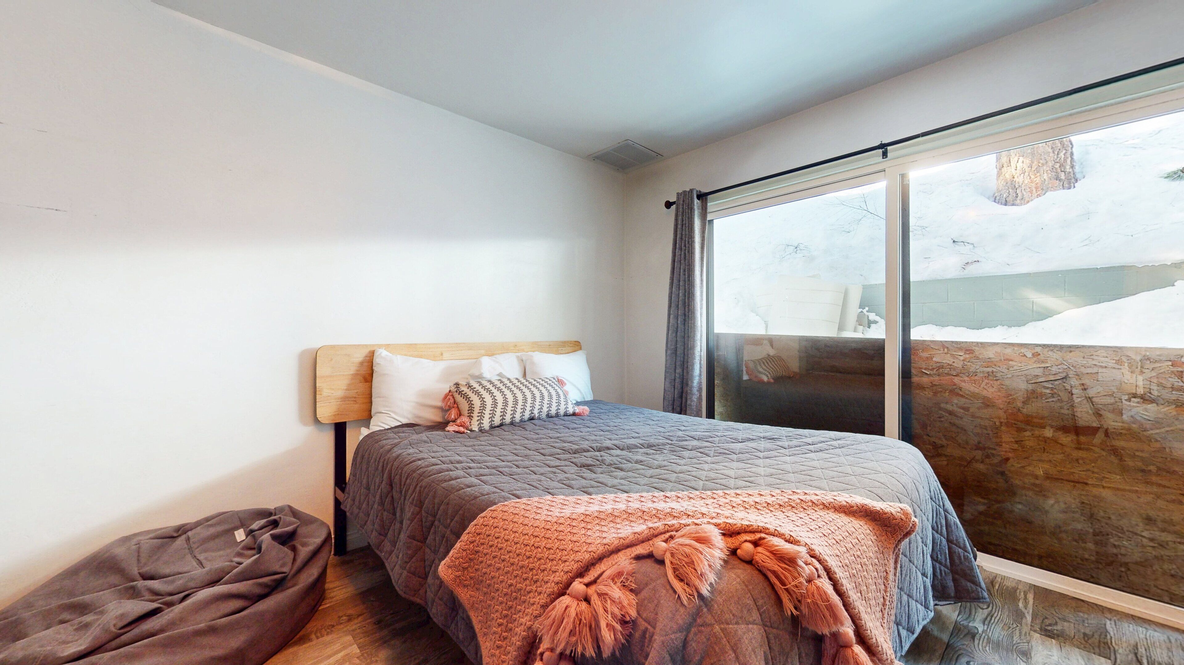 A bedroom with a bed, a wooden headboard, a window with a view of a snowy landscape, and a wooden floor.
