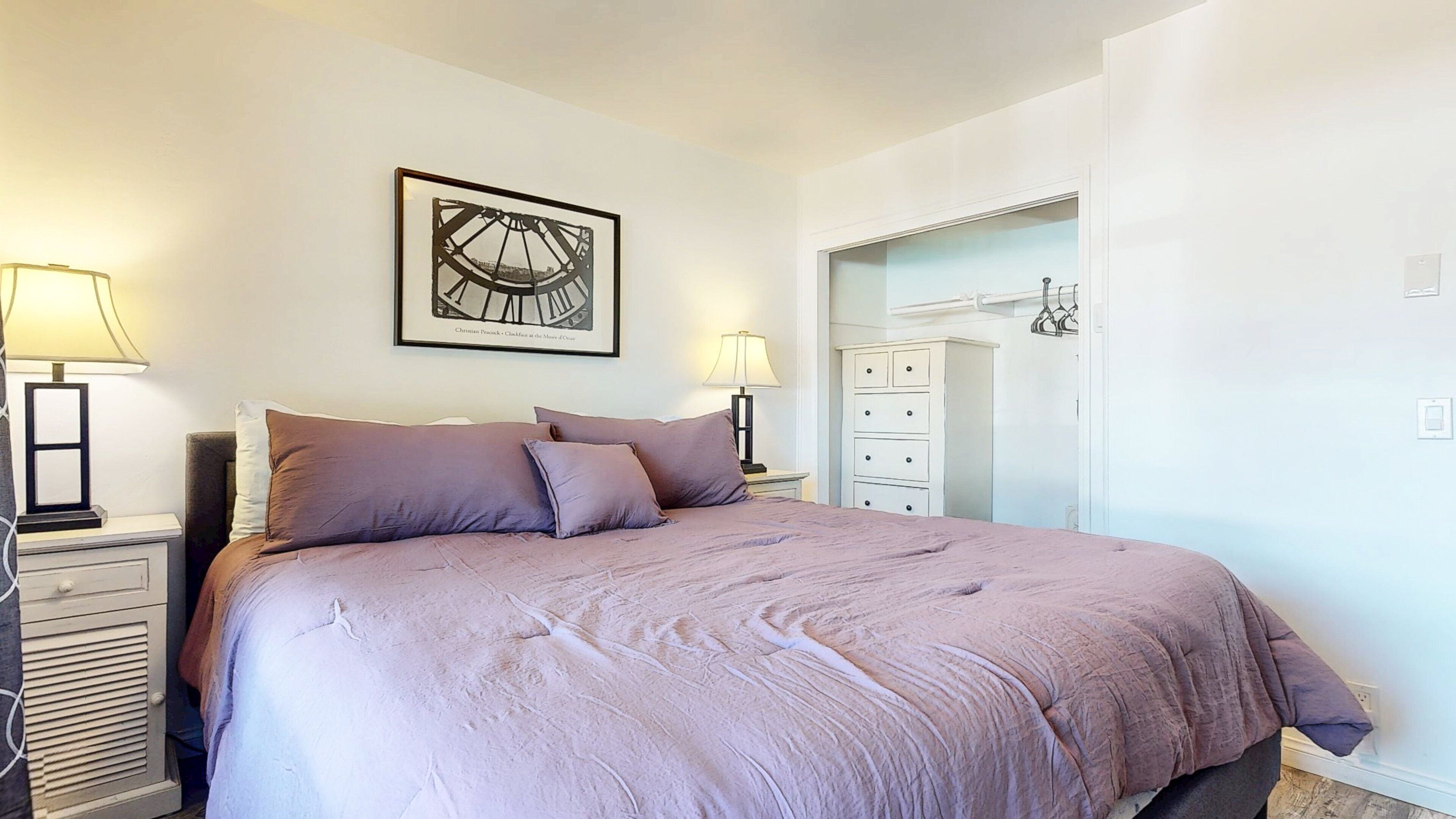 A bedroom with a large bed, two bedside tables with lamps, a dresser, and a framed picture on the wall.