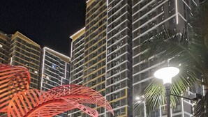 Vista nocturna de una piscina de hotel con sillones de descanso, una estructura roja decorativa y edificios altos al fondo.