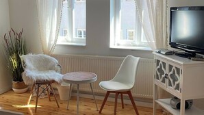 A living room with a wooden floor, a white sofa, a white chair, a small round table, a TV on a white cabinet, and a large window with sheer curtains. - Charming half-timbered house in the heart of the city "Fachwerkstube" 3 Bedroom Apartment (Witzenhausen)