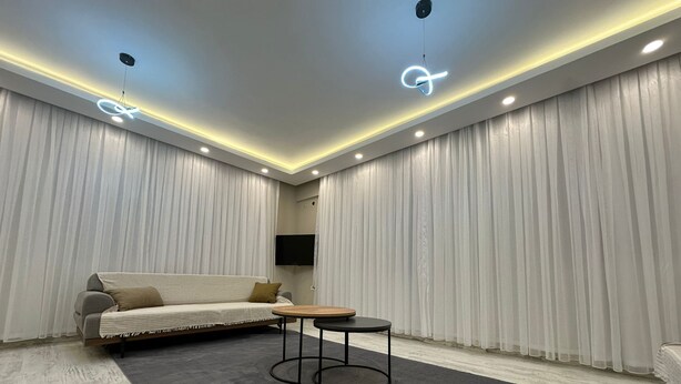 Una sala de estar moderna con un sofá, una mesa de centro y un televisor de pantalla plana frente a una pared con cortinas translúcidas.