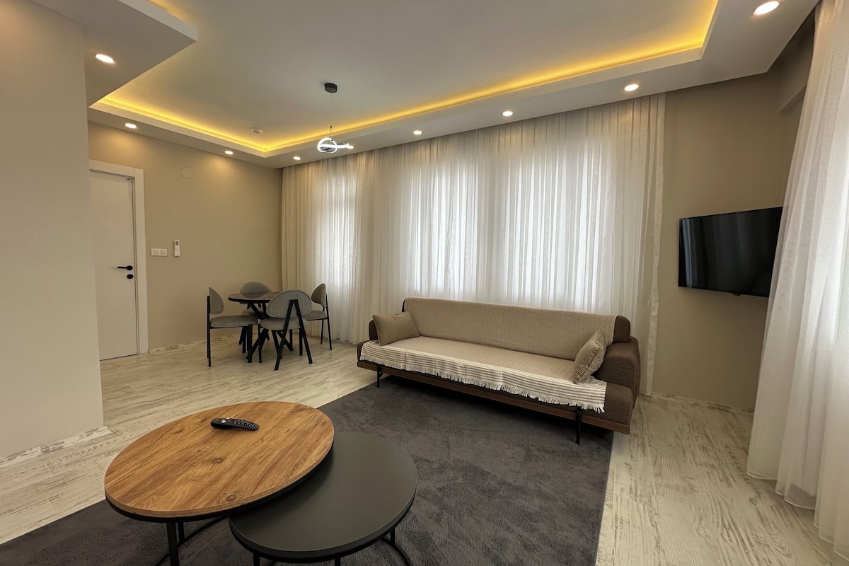 Uma sala de estar moderna com um sofá, uma mesa de jantar com cadeiras, uma TV fixada na parede e um teto com iluminação embutida.
