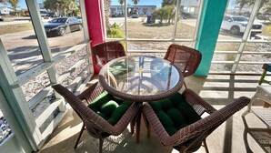 Uma área de estar coberta ao ar livre, com cadeiras de vime e uma mesa de vidro, com vista para um estacionamento e edifícios.