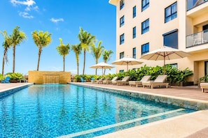 A swimming pool with lounge chairs and umbrellas, surrounded by palm trees and a building with multiple windows.
