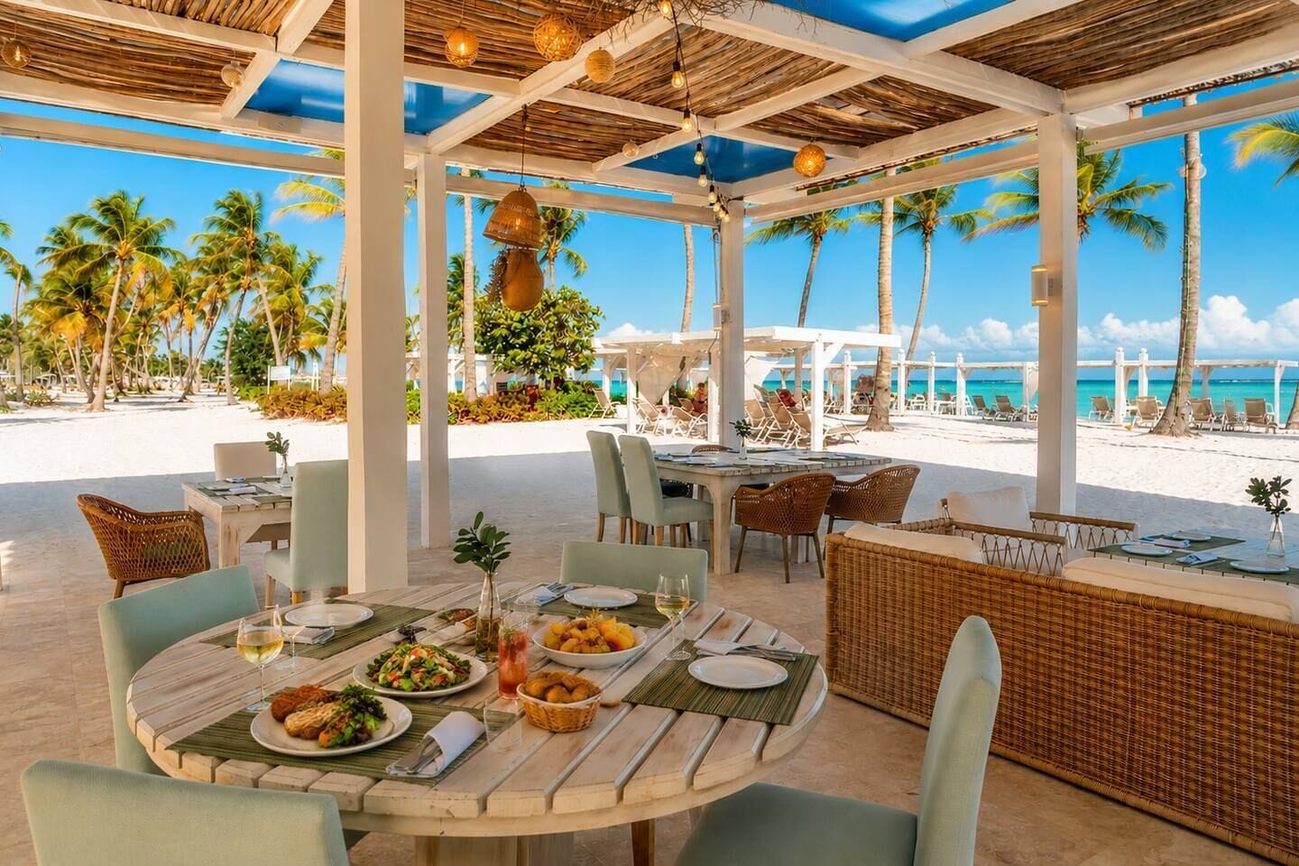 Un'area pranzo all'aperto coperta con tavoli e sedie, vista sulla spiaggia e palme.