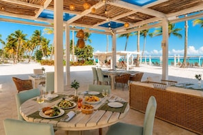 A covered outdoor dining area with tables and chairs, a view of the beach, and palm trees.