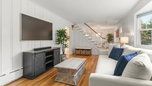 Una sala de estar moderna con un televisor de pantalla plana, un mueble de entretenimiento gris, una mesa de centro de mimbre, un sofá blanco con cojines azules y una escalera que conduce a un piso superior.