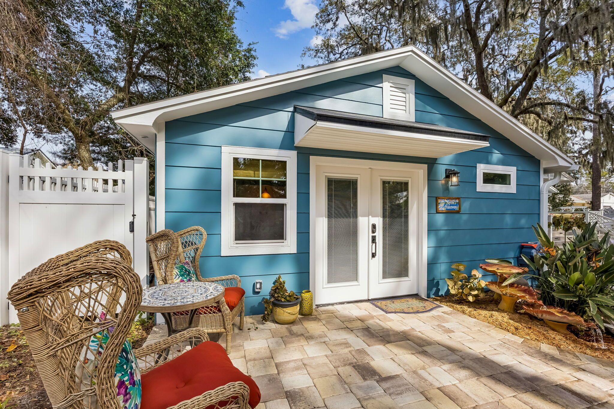 Una pequeña casa con fachada azul, una puerta blanca y un patio con muebles de mimbre y plantas en macetas.