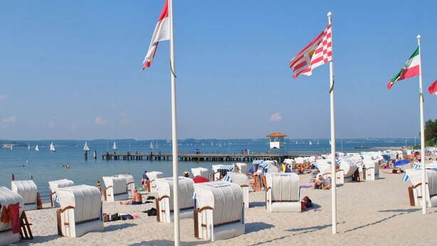 A beach with rows of white wicker lounge chairs, a pier in the background, and flags on poles. - At the small mill pond - central quiet location with parking and bicycles (Flensburg)