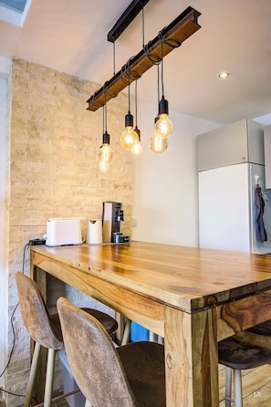 Eine moderne Küche mit einem Holztisch, hängenden Lampen, einer Kaffeemaschine und einem Kühlschrank.
