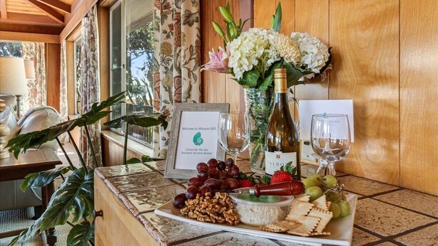 Une table avec une bouteille de vin, des verres, des raisins, des noix et des crackers, ainsi qu’un panneau d’accueil encadré et un arrangement floral.