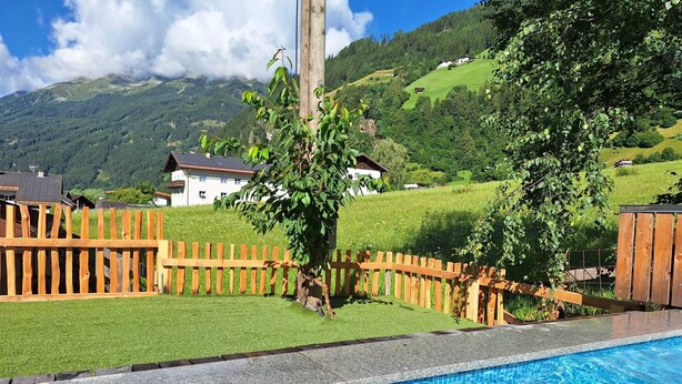 A swimming pool with a wooden fence and a view of a green landscape with houses in the background. - Dorf Apart & Dorf Cafe Appartements (Neustift Im Stubaital)