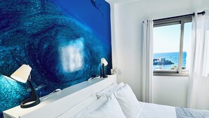 Une chambre d’hôtel avec un grand lit, du linge de lit blanc, une table de chevet avec une lampe, un téléviseur fixé au mur qui diffuse une image d’un océan bleu, et une fenêtre donnant sur la mer et un bâtiment situé au loin.