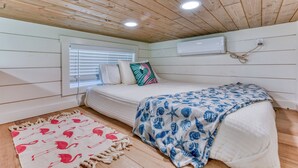 A cozy bedroom with a wooden ceiling, a bed with a blue and white blanket, a red and white rug, and a window with blinds. - Tiny Islands Resort – Aruba | Tropical Tiny Home with Loft, Fire Pit & Style (Blanco)