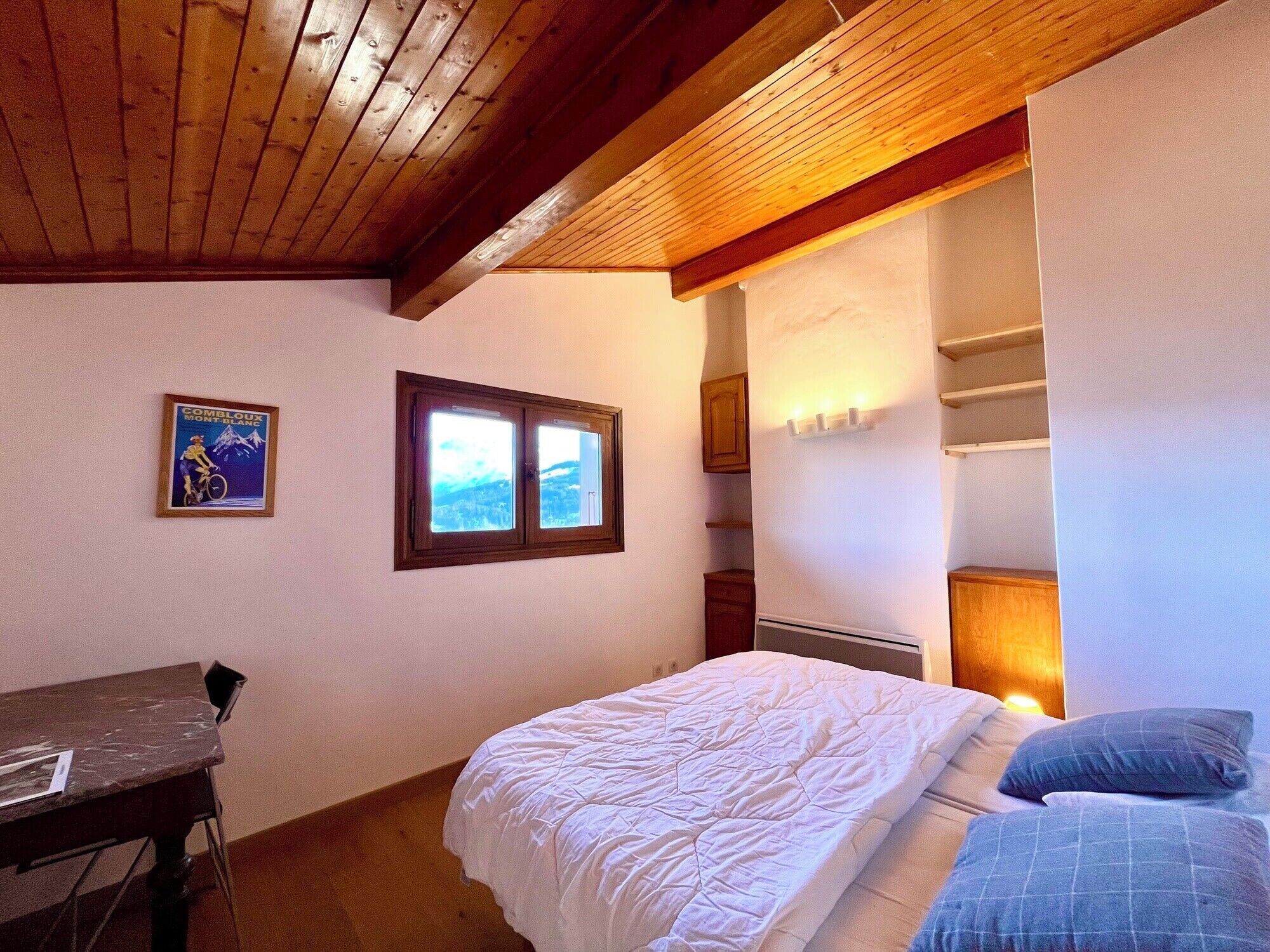 A bedroom with a bed, a desk, a window, and a wall-mounted shelf.
