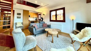 En moderne stue med en grå sofa, et lille rundt bord og en hvid, lodden stol. Et køkken er synligt i baggrunden med træskabe og et vindue.