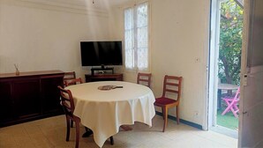 Uma sala de jantar com uma mesa redonda, cadeiras, uma televisão e uma janela com vista para o verde.
