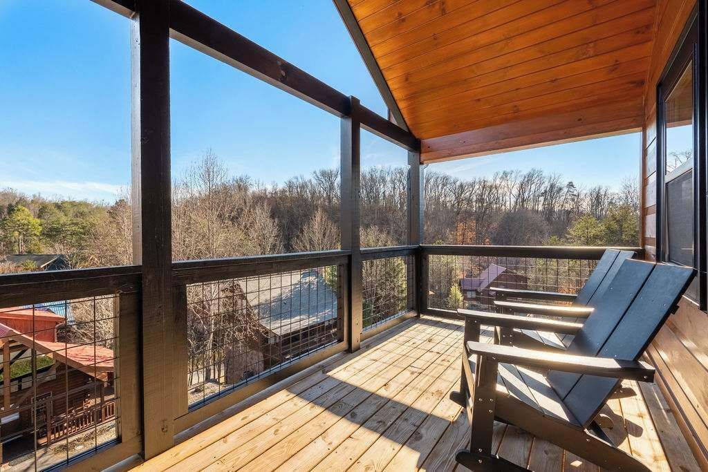 A wooden deck with a metal railing, two Adirondack chairs, and a view of a forested area.