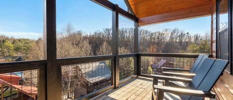 A wooden deck with a metal railing, two Adirondack chairs, and a view of a forested area.