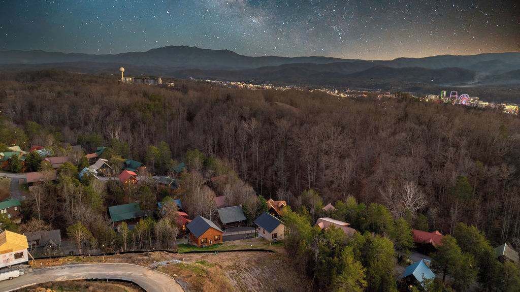 Área residencial com casas cercadas por floresta, uma estrada sinuosa e uma vista distante de uma cidade com edifícios iluminados e uma roda gigante.