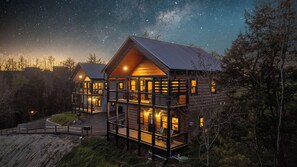 A two-story log cabin with a balcony and a deck, surrounded by trees and a starry night sky. - Modern Pigeon Forge Cabin | Hot Tub, Game Loft (Pigeon Forge)