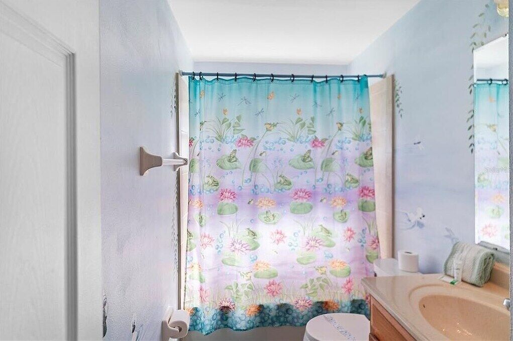 Un baño con cortina de ducha estampada con motivos florales, un inodoro blanco, un lavabo con espejo y un perchero para toallas.