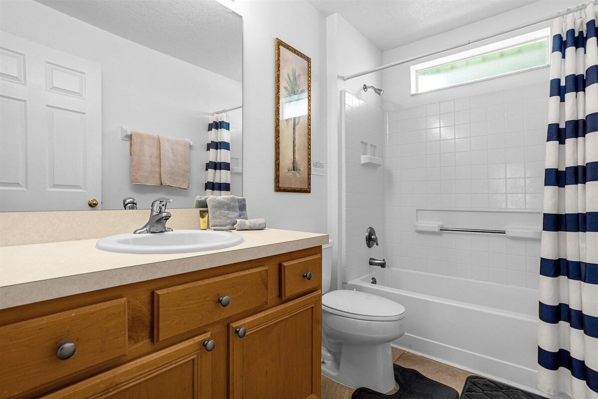Ein Badezimmer mit weißem Waschbecken, Holz-Waschtisch, einem gerahmten Bild an der Wand, einer weißen Toilette, einer Badewanne und einer Dusche mit blau-weiß gestreiftem Vorhang.