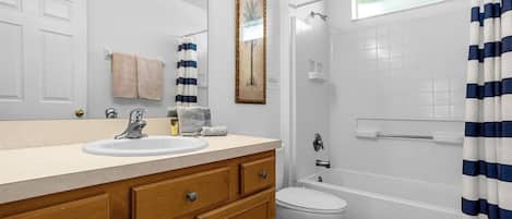 Ein Badezimmer mit weißem Waschbecken, Holz-Waschtisch, einem gerahmten Bild an der Wand, einer weißen Toilette, einer Badewanne und einer Dusche mit blau-weiß gestreiftem Vorhang.