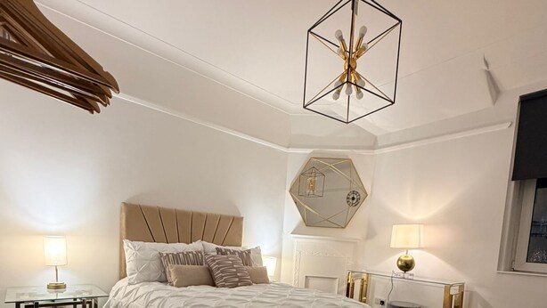 Una camera da letto moderna con un grande letto, una lampada a soffitto geometrica, uno specchio esagonale e un piccolo comodino con una lampada.