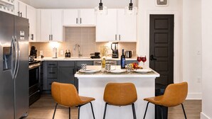 Cocina moderna con armarios blancos, un refrigerador de acero inoxidable, una isla central con taburetes altos e iluminación colgante.