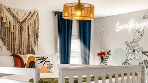 Una cuna blanca con barandilla blanca, un adorno de macramé colgando en la pared, una lámpara fijada a la pared y una pared con diseño floral y un letrero de neón.