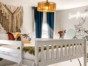 Una cuna blanca con barandilla blanca, un adorno de macramé colgando en la pared, una lámpara fijada a la pared y una pared con diseño floral y un letrero de neón.