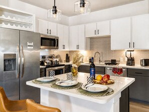 A modern kitchen with stainless steel appliances, white cabinetry, and a central island with a water dispenser and a bottle of wine. - Modern Apt Minutes to Universal (Frisco)