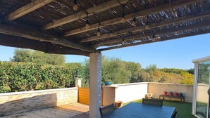 A covered outdoor patio with a table and chairs, a wooden deck, and a view of greenery and hills in the background. - "L'Olivier" Vacation Rental - Avignon, Nîmes, Uzès, St. Rémy (Aramon)