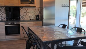 A modern kitchen with wooden cabinets, a stainless steel refrigerator, a built-in oven, and a dining table with chairs. - "L'Olivier" Vacation Rental - Avignon, Nîmes, Uzès, St. Rémy (Aramon)