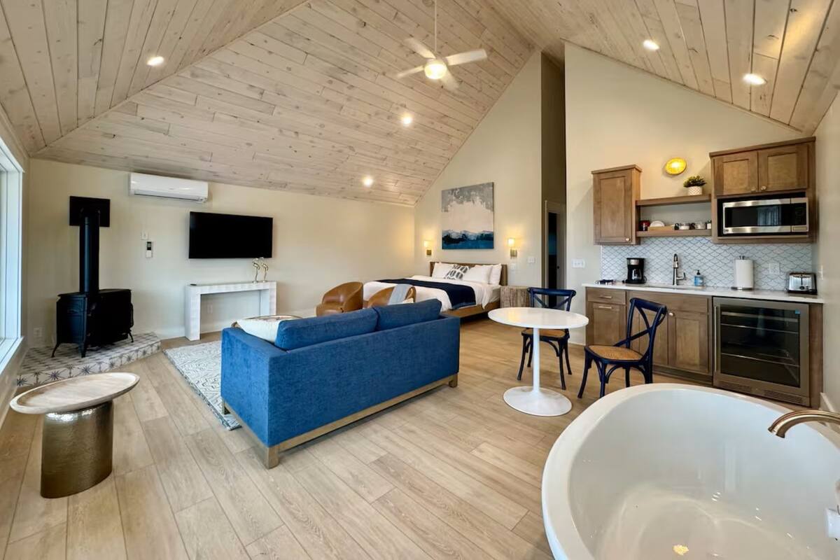 A modern interior with a loft-style ceiling, a cozy seating area with a blue sofa, a dining table, a kitchen with wooden cabinets, and a large bathtub.