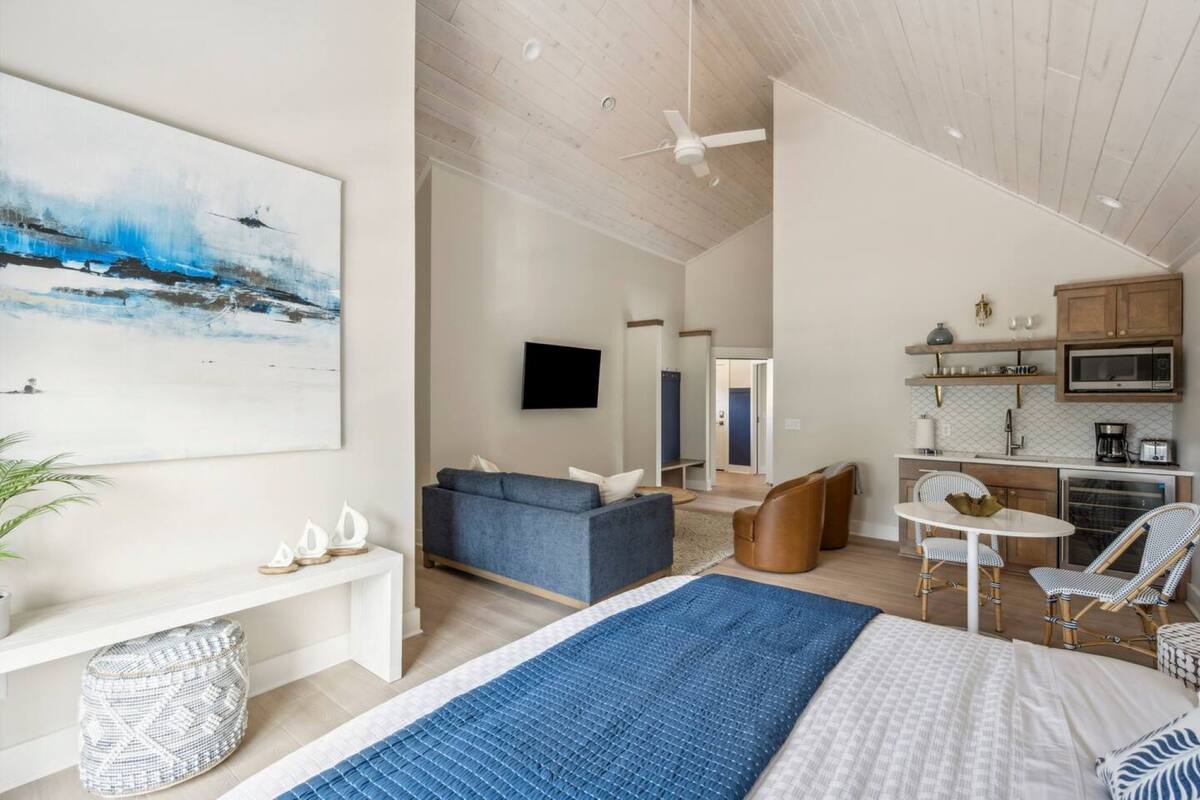 A modern hotel room with a large bed, a blue area rug, a sofa, a dining table with chairs, a kitchen area, and a flat-screen TV.
