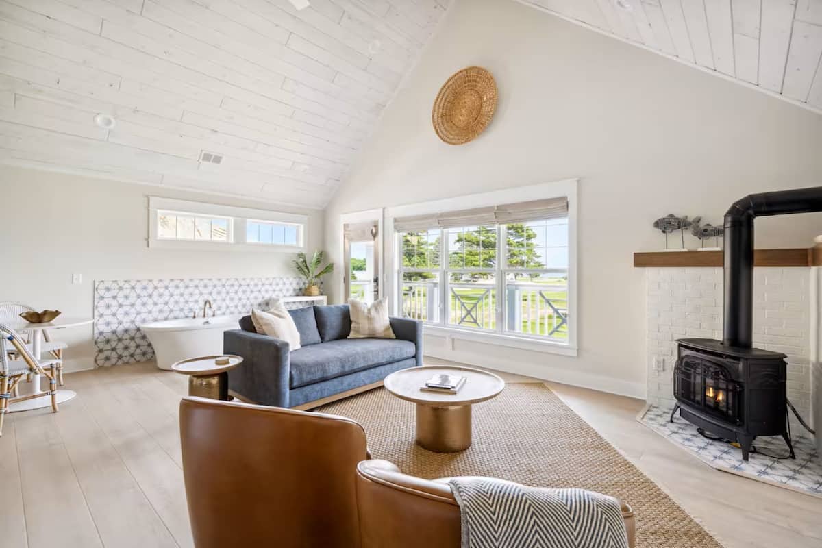 A spacious living room with a sofa, armchair, and coffee table, a fireplace, and large windows offering a view of the outdoors.