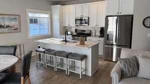 A modern kitchen with white cabinets, stainless steel appliances, a central island with bar stools, and a dining area with a round table and chairs. - Harbor Splash - Steps to Indoor Pool (South Haven)