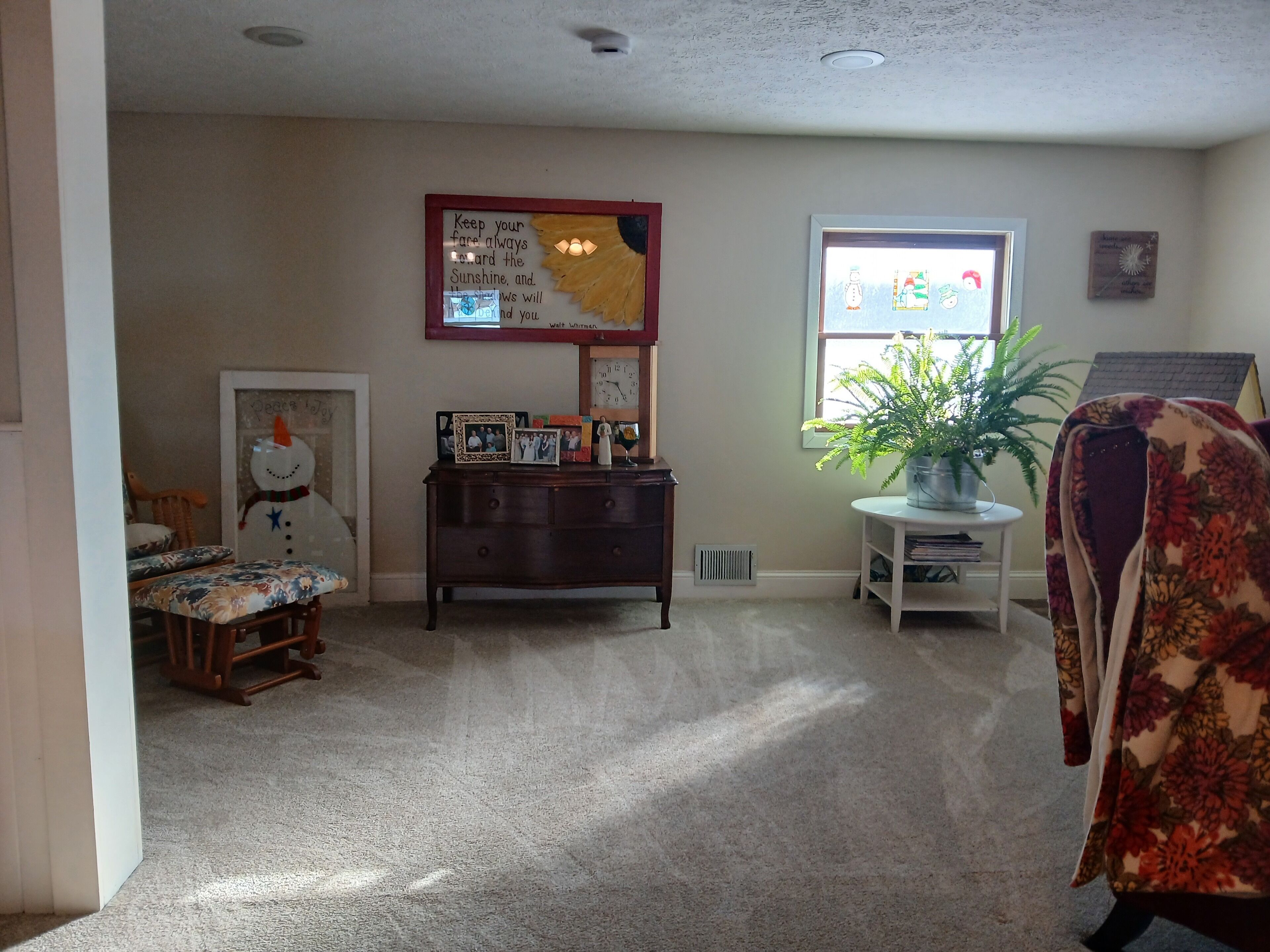 A cozy living room with a wooden dresser, a floral chair, a small table with a plant, a framed picture, and a wall clock.
